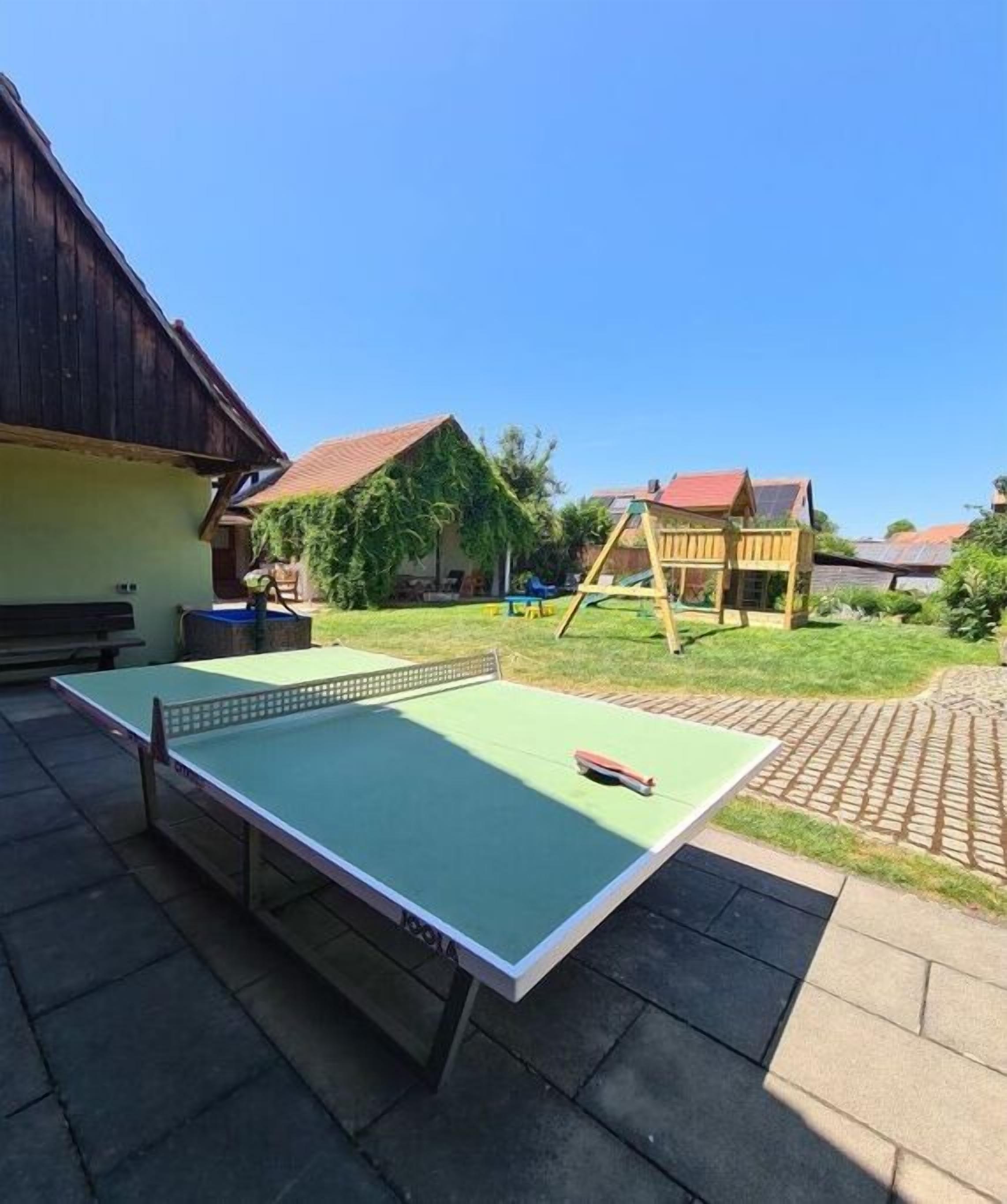 Une table de ping-pong installée à l’extérieur, dans une cour, avec une structure de jeu en bois en arrière-plan.