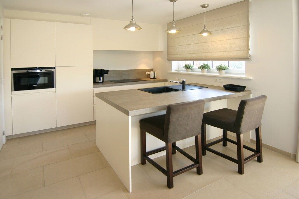 Een moderne keuken met een centraal eiland, twee barkrukken, een inbouwoven en hanglampen.