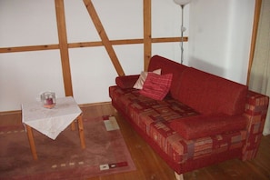 Ein rotes Sofa mit gemusterten Kissen, ein kleiner Tisch mit Glas und Kerze sowie ein Holzboden.