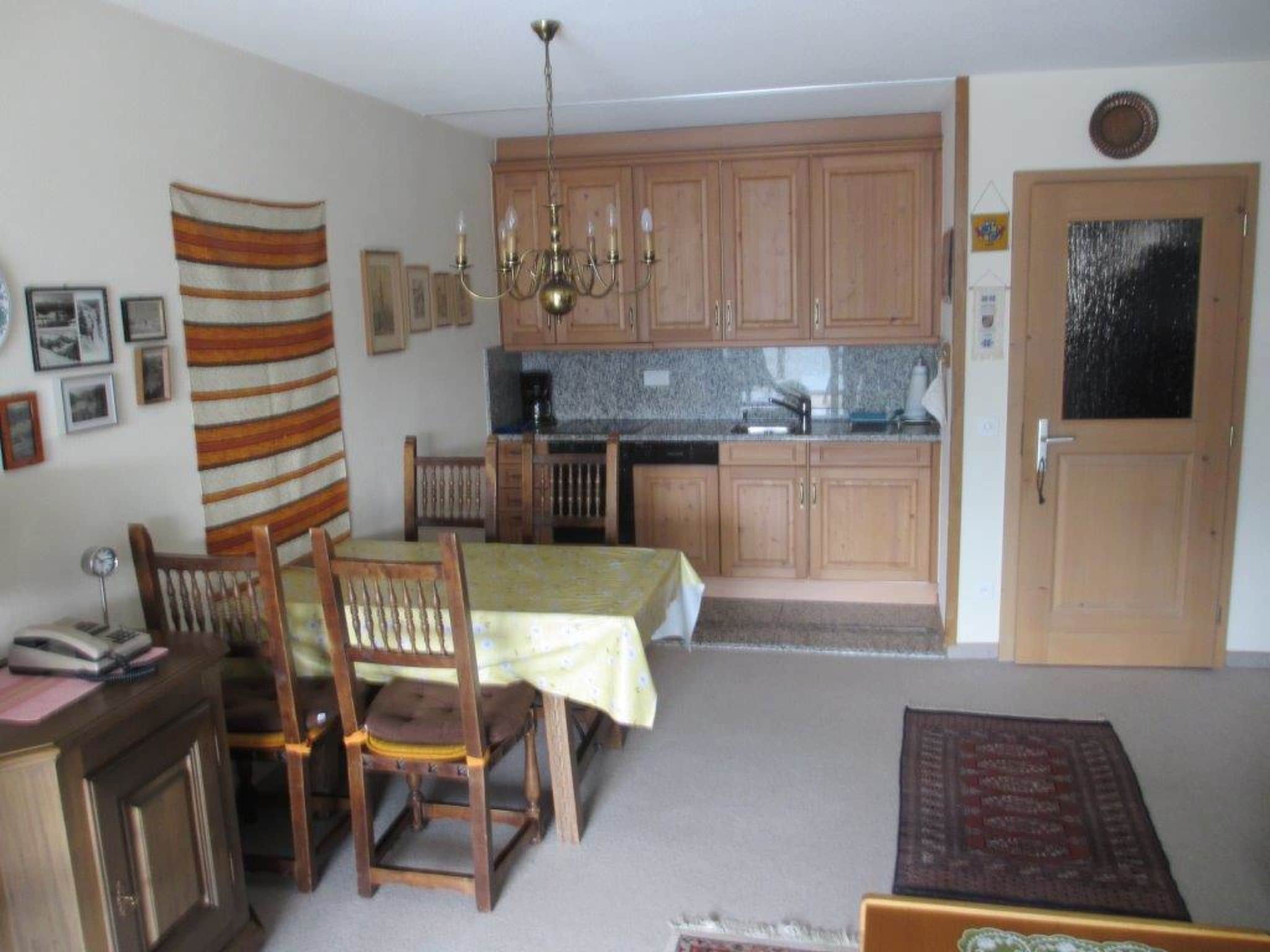 A compact kitchen with wooden cabinets, a dining table with chairs, a chandelier, and a door with a frosted glass panel.