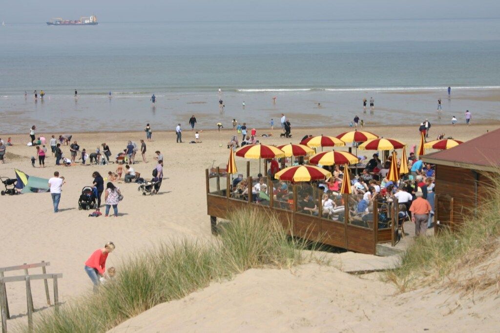Ein Strand mit einer Holzkonstruktion, roten und gelben Sonnenschirmen, Personen die spazieren gehen und einem Schiff in der Ferne.