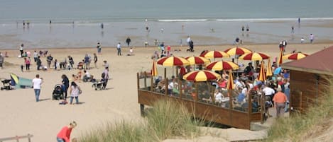 Een strand met een houten constructie, rode en gele parasols, mensen die wandelen en een schip in de verte.