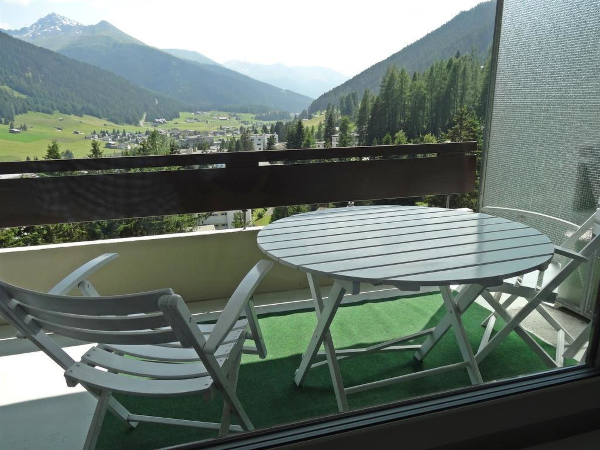 A balcony with a round table and two chairs, overlooking a valley with mountains and a town.