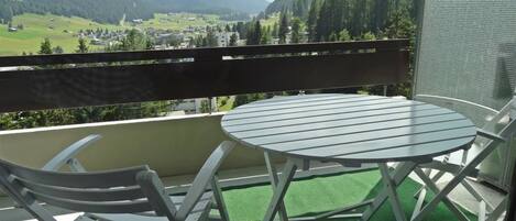 A balcony with a round table and two chairs, overlooking a valley with mountains and a town.