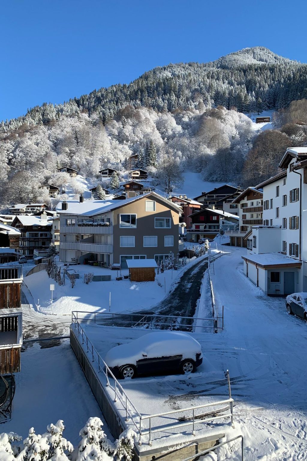 A snow-covered residential area with houses, a car, and a mountain in the background.