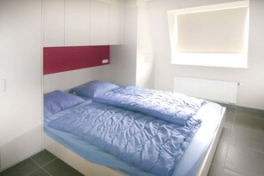 A single bed with a headboard, a window with blinds, and a radiator on the wall. - Holiday apartment with 1 bedroom (Cadzand)
