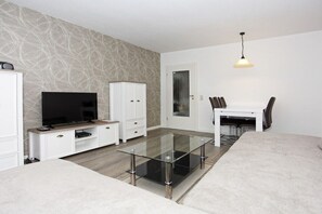 A modern living room with a glass coffee table, white cabinetry, a flat-screen TV, and a dining area with chairs. - Holiday apartment with 1 bedroom (Borkum)