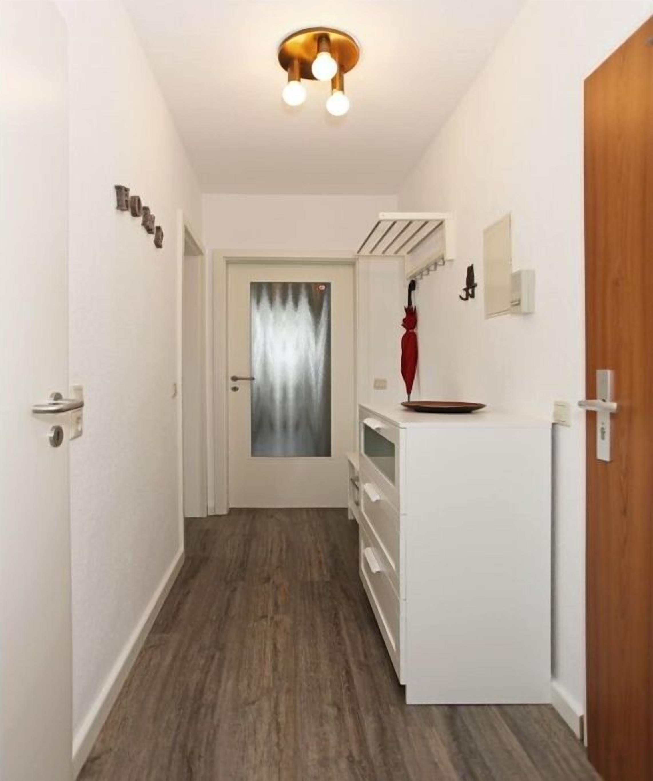 Un pasillo estrecho con piso de madera, una puerta blanca al final y un armario pequeño con un paraguas rojo encima.