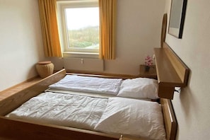Ein Einzelbett mit Holzrahmen und weißer Bettwäsche, ein Nachttisch mit Lampe, ein Fenster mit Vorhängen sowie ein kleiner Topfpflanze.