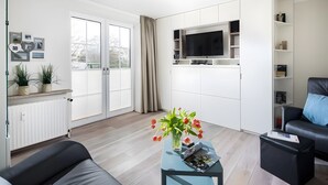 A modern living room with a sofa, a coffee table with a vase of flowers, a television mounted on the wall, and a sliding glass door leading to a balcony. - Apartment “Seestern” with Balcony and Wi-Fi (Norderney)