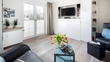 Ein modernes Wohnzimmer mit einer Couch, einem Couchtisch mit Blumen in einer Vase, einem an der Wand montierten Fernseher und einer Glastür, die auf einen Balkon führt.