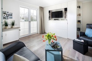 Ein modernes Wohnzimmer mit einer Couch, einem Couchtisch mit Blumen in einer Vase, einem an der Wand montierten Fernseher und einer Glastür, die auf einen Balkon führt.