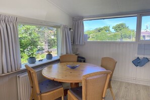 A dining area with a round table and four chairs, a window with curtains, and a view of greenery outside. - Previnaireweg 46 & 48 (Callantsoog)