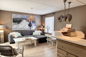 A modern living room with a grey sofa, white coffee table, wicker chairs, a large ocean-themed painting, and a bar area with a beverage dispenser. - Holiday apartment with 1 bedroom (Callantsoog)