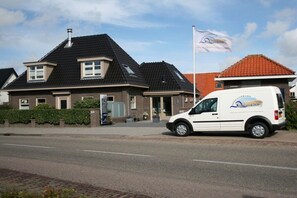A building with a black roof, a white van parked in front, and a flag on a flagpole. - Holiday apartment with 1 bedroom (Callantsoog)