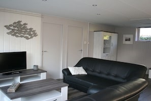 A living room with a black leather sofa, a flat-screen TV on a stand, a small table with a glass container, and a white cabinet. - Bungalow with 1 bedroom (Callantsoog)