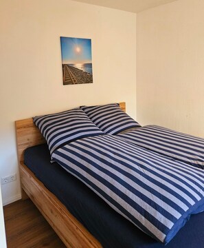 A bed with a wooden frame and striped bedding, a wall-mounted picture of a sunset, and a power outlet on the wall.