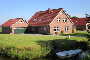 A two-story brick house with a red roof, a garage, and a small boat docked by a calm water body. - Biesterfeld holiday apartment (Bedekaspeler Marsch  \/ Südbrookmerland)