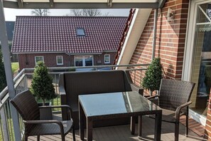 A balcony with wicker furniture and a glass table, overlooking a red-tiled roof house. - Biesterfeld holiday apartment (Bedekaspeler Marsch  \/ Südbrookmerland)