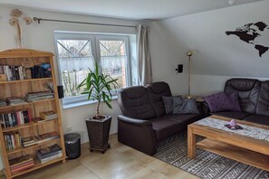 Ein Wohnzimmer mit einer Couch, einem Couchtisch, einem Bücherregal, einer Topfpflanze und einem Fenster mit Vorhängen.