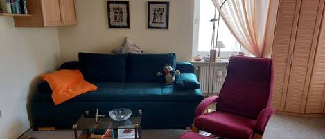 Una sala de estar con un sofá azul oscuro, un sillón mecededor rojo, una mesa de centro de vidrio y una estantería.