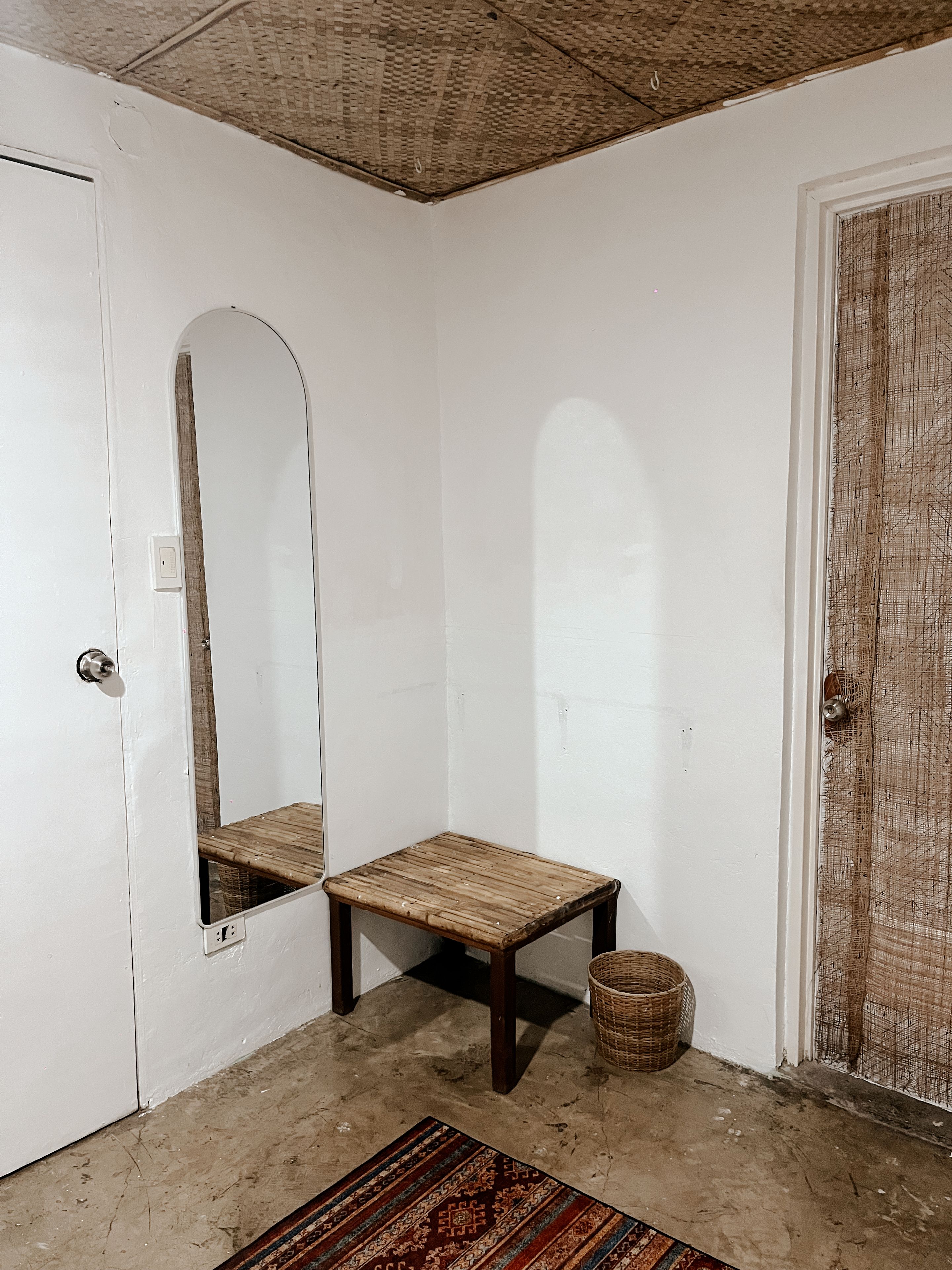A room with a white door, a small wooden bench, a woven basket, and a patterned rug.