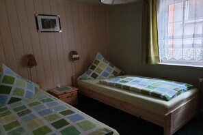 Ein Zimmer mit zwei hölzernen Betten, einem kleinen Nachttisch, einem an der Wand hängenden Bild und einem Fenster mit Vorhang.