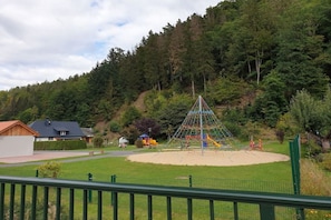A playground with climbing structures and slides, surrounded by a fenced area with a grassy field and a house in the background. - Schiel Country House (Rohrbach)