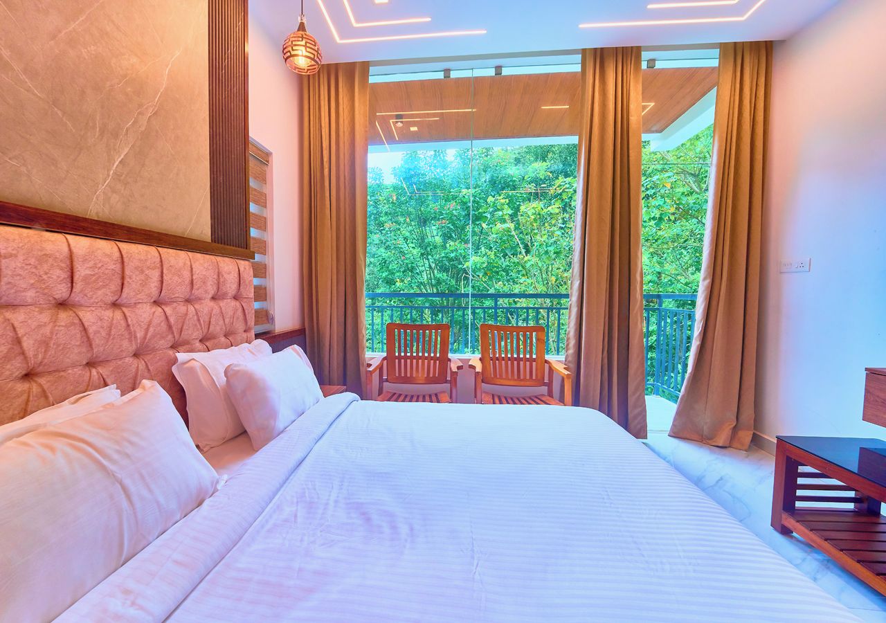 Habitación de hotel con una cama grande, cabecera de madera, balcón con sillas y vista a zonas verdes.