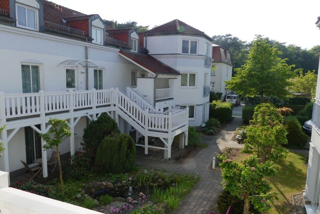 Ein mehrstöckiges Wohngebäude mit einer überdachten Außenfläche, einer Treppe und einem Garten mit verschiedenen Pflanzen und einem kleinen Teich.