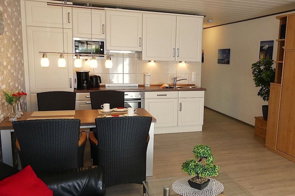 A compact kitchen with white cabinets, a dining area with wicker chairs, and a small potted plant.