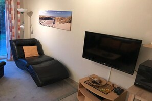 Un salon avec un canapé en cuir noir, un téléviseur à écran plat fixé au mur, une petite table d’appoint en bois avec un plateau, et un tableau représentant une scène de plage.