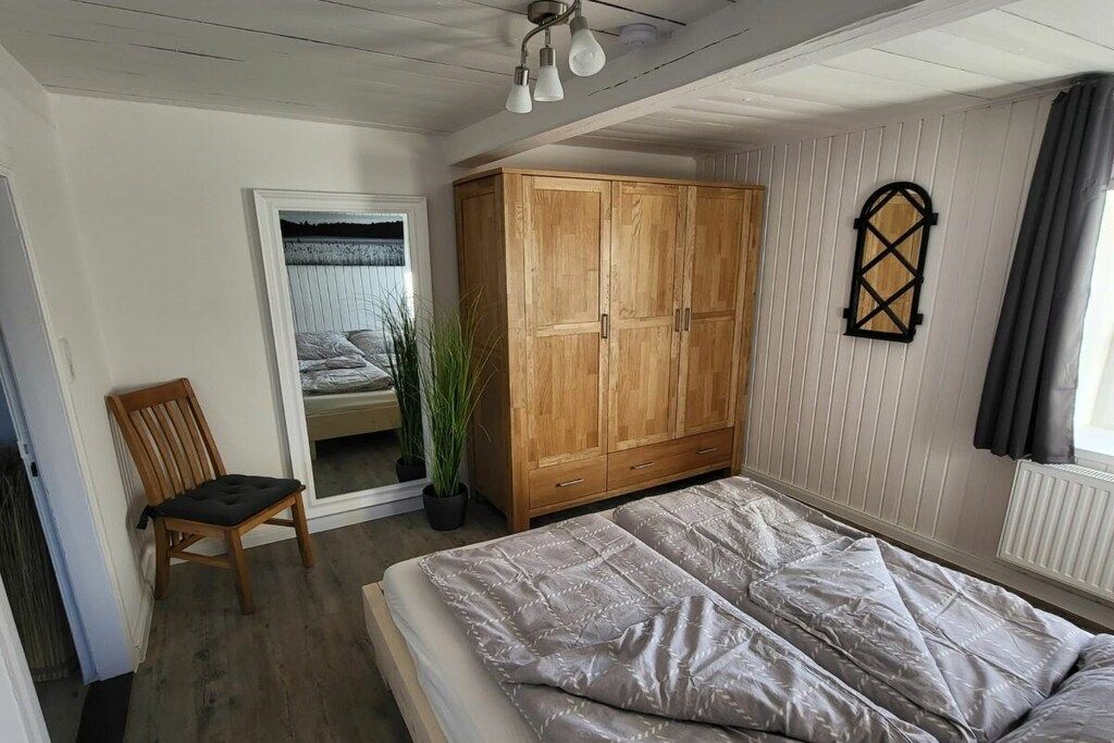 Een slaapkamer met een bed, een houten garderobe, een stoel, een kamerplant en een raam met gordijnen.
