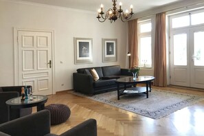 Ein Wohnzimmer mit Sofa, Couchtisch und Sesseln, einer Kristallleuchtre, gerahmten Bildern an der Wand und großen Fenstern mit Vorhängen.
