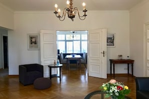 A spacious living room with a chandelier, a dining area with a table and chairs, and a door leading to another room. - Villa Achterkerke / 1st floor (Seebad Heringsdorf)