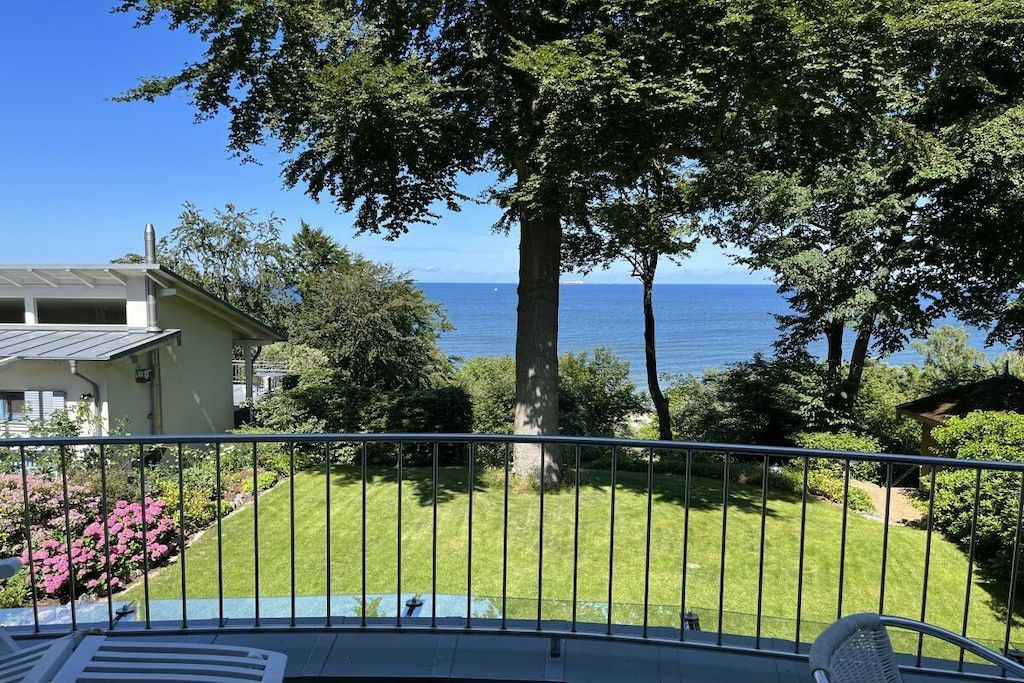 En balkon med udsigt over havet, en græsplæne og et hus i baggrunden.