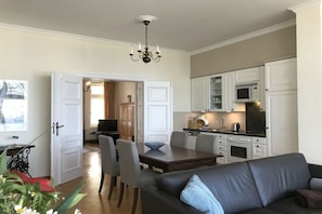 A modern kitchen with white cabinets, a dining area with a dark wooden table and grey chairs, and a black leather sofa with a blue pillow. - Villa Achterkerke / 1st floor (Seebad Heringsdorf)