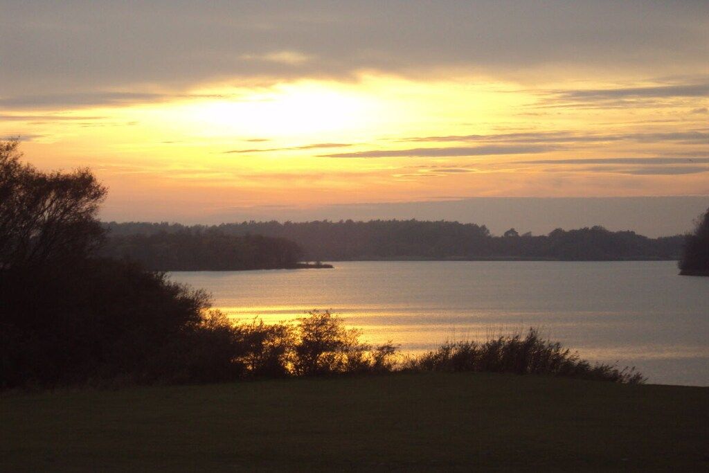 Ein See bei Sonnenuntergang, bewölkter Himmel, umgeben von Bäumen und Vegetation.