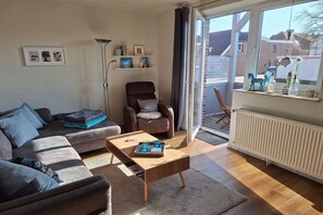 A modern living room with a sofa, armchair, coffee table, and a view through a glass door. - Holiday apartment with 1 bedroom (Hooksiel)