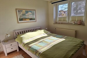 Een slaapkamer met een bed, nachtkastje, raam met jaloezieën en een ingelijst beeld aan de muur.