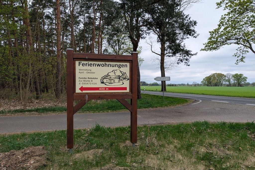 En skylt med text och en pil som visar riktning, placerad vid sidan av en väg med en gräsyta och träd i bakgrunden.