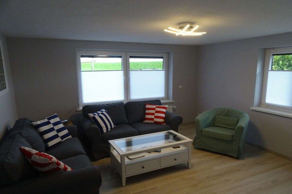 Ein Wohnzimmer mit einer Sofagarnitur, einem Couchtisch und einem Sessel. Zwei Fenster mit Jalousien und eine Deckenleuchte sind vorhanden.