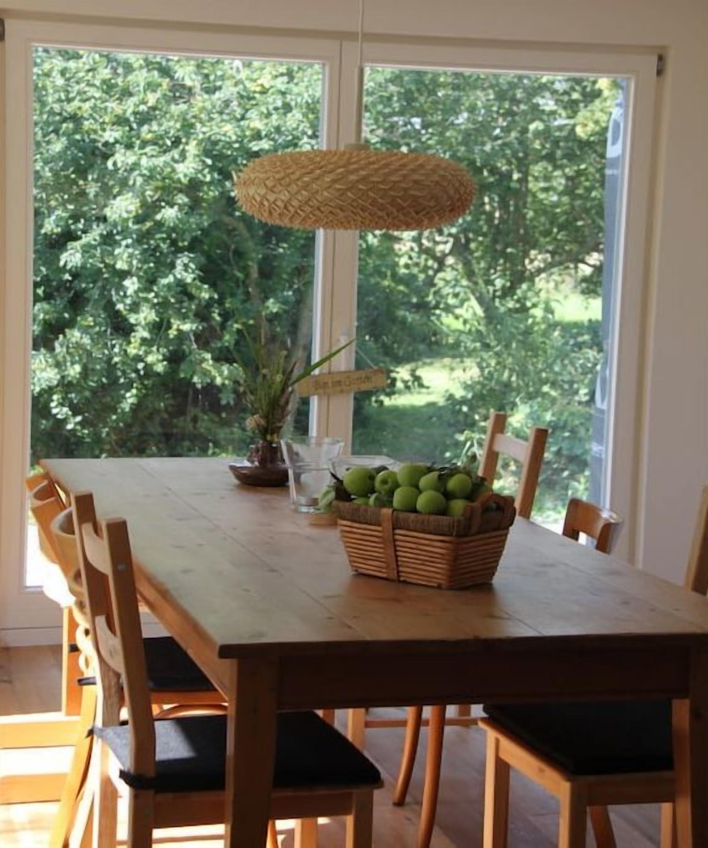 Uma mesa de jantar de madeira com cadeiras, uma luminária pendente de palha e uma cesta de frutas.