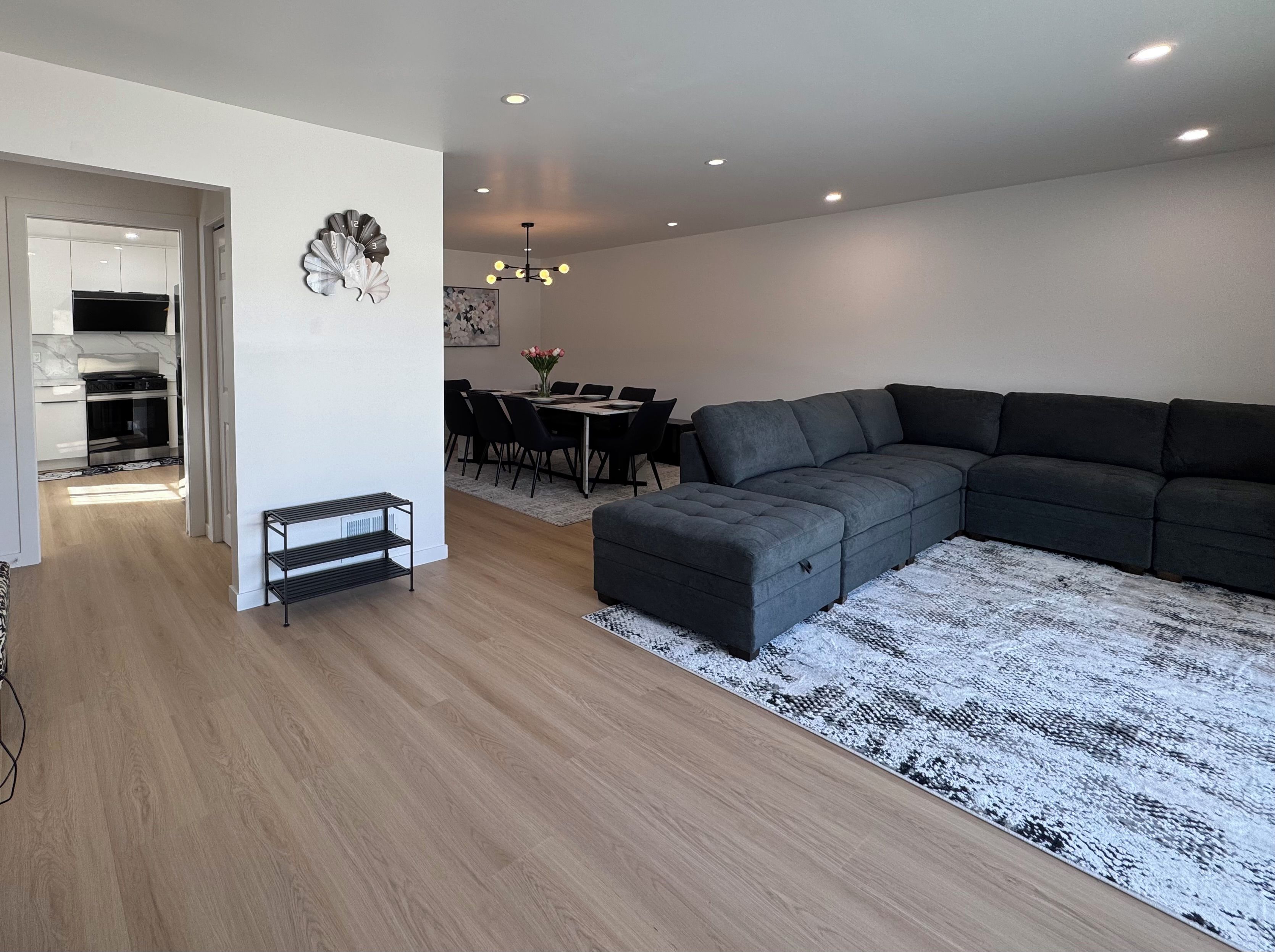 Un salon moderne avec un canapé d’angle, une table à manger avec des chaises, un lustre et une cuisine en arrière-plan.