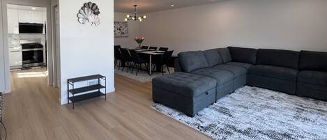 Un salon moderne avec un canapé d’angle, une table à manger avec des chaises, un lustre et une cuisine en arrière-plan.