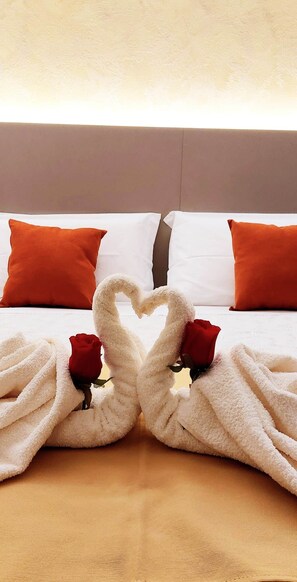 Een bed met witte en oranje kussens, een handdoekarrangement in de vorm van een hart, en twee rode rozen.