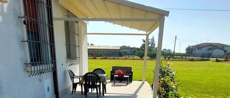 A covered outdoor seating area with a patio table and chairs, a small garden with potted plants, and a view of a green field.