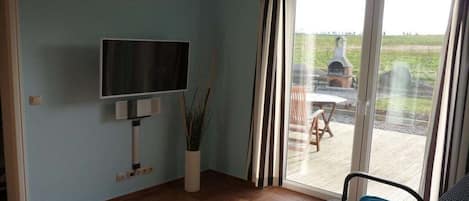 A room with a television mounted on the wall, a potted plant, a sliding glass door leading to an outdoor area with a barbecue grill, and a chair.
