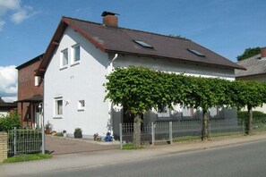 Ein zweistöckiges Haus mit weißer Fassade, braunem Dach und einer überdachten Veranda mit Grünpflanzen.