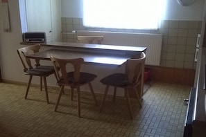 Et køkken med flisegulv, et træbord, fire stole, et vindue med persienner, en radiator og en lille elektronisk enhed på bordet.